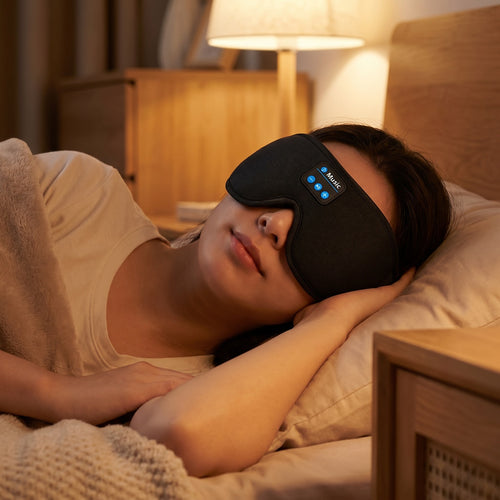 A woman lying in bed wearing a black sleep mask with blue LED indicator lights, in a warmly lit bedroom setting.